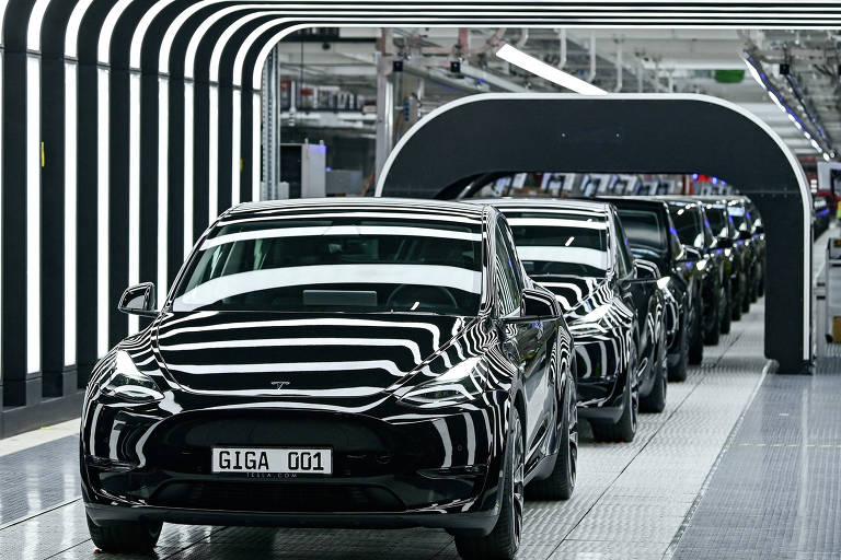 A imagem mostra uma linha de montagem de carros Tesla, com vários veículos pretos alinhados em uma estrutura moderna e iluminada. O ambiente é caracterizado por luzes em formato de faixas que criam um efeito visual interessante. Os carros estão em uma superfície de piso claro, e a placa de um dos veículos exibe a inscrição 'GIGA G01'.