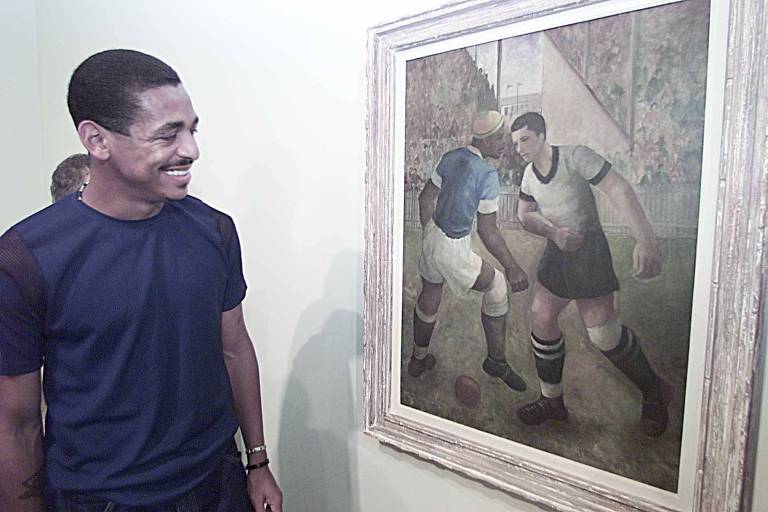 Um homem sorridente está em pé ao lado de uma pintura que retrata dois jogadores de futebol em um campo. Um dos jogadores está vestindo uma camisa azul e calças brancas, enquanto o outro usa uma camisa branca e calças escuras. A pintura é emoldurada e há uma multidão visível ao fundo, sugerindo um ambiente de jogo. O homem está usando uma camiseta escura e parece apreciar a obra de arte.