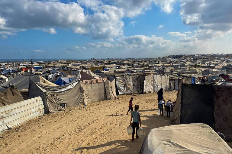 A imagem mostra um grande campo de refugiados, com várias tendas dispostas em uma área de areia. No fundo, há um céu parcialmente nublado e uma linha de horizonte que sugere a presença de água. Algumas pessoas estão caminhando pelo campo, carregando objetos. As tendas são de diferentes tamanhos e cores, predominantemente em tons de cinza e bege.