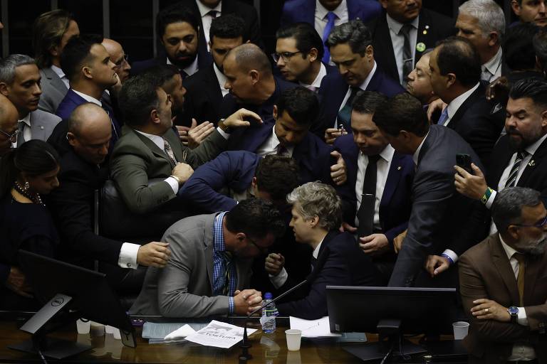 A imagem mostra um grupo de pessoas em um ambiente de debate, aparentemente no Congresso. Há uma agitação visível, com várias pessoas se aglomerando e interagindo de forma intensa. Algumas estão em pé, enquanto outras estão sentadas, e todos parecem estar envolvidos em uma discussão acalorada. O ambiente é formal, com mesas e computadores visíveis ao fundo.