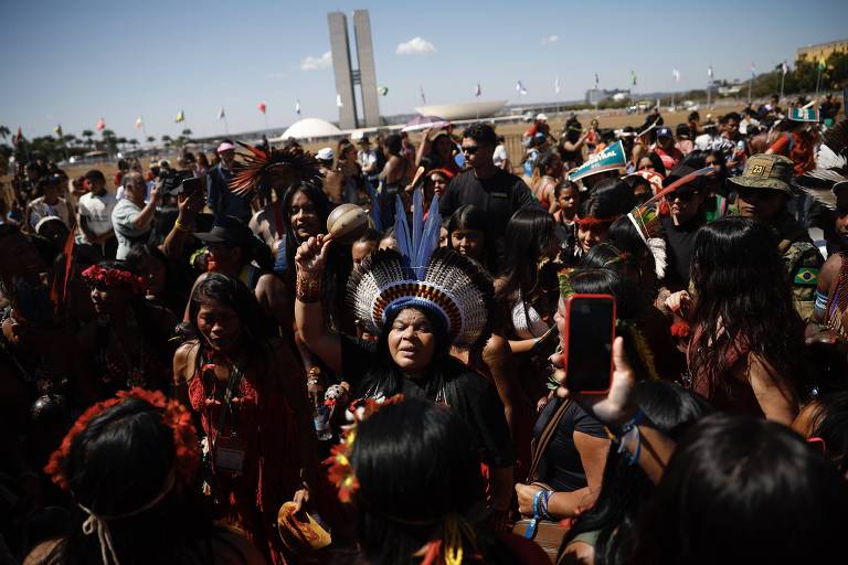 A imagem mostra um grande grupo de pessoas, predominantemente indígenas, reunidas em um espaço aberto. Algumas pessoas estão vestindo trajes tradicionais e adornos, como penas e colares. No centro, uma mulher com um cocar de penas levanta a mão, parecendo expressar emoção ou fazer um discurso. Ao fundo, há bandeiras e estruturas que sugerem um evento ou manifestação. O céu está claro e há algumas nuvens.