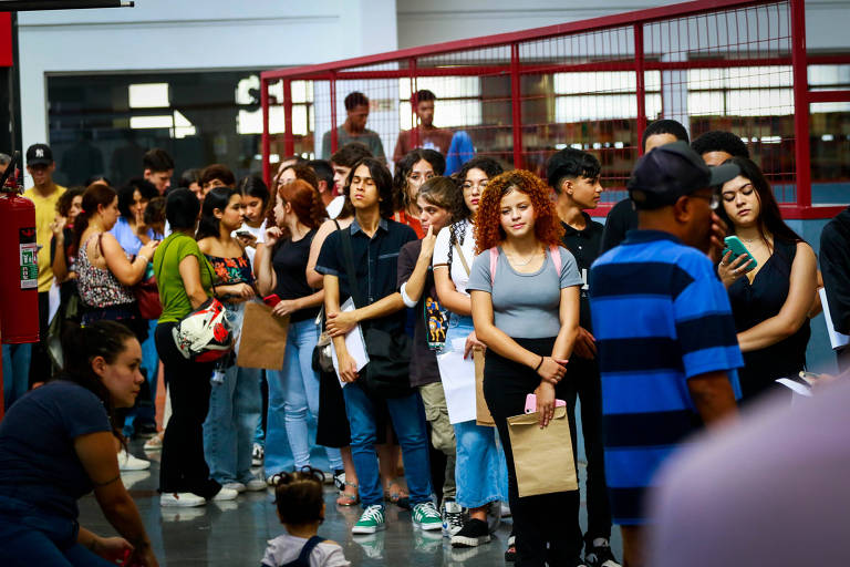 A imagem mostra uma fila de pessoas em um ambiente interno, possivelmente em um evento ou local de inscrição. As pessoas estão em pé, algumas segurando documentos ou pastas. O ambiente é bem iluminado, com uma estrutura metálica ao fundo. Há uma diversidade de estilos de roupas entre os participantes, que parecem ser jovens. No primeiro plano, uma mulher com cabelo cacheado e uma blusa clara está em destaque, enquanto outras pessoas estão visíveis ao fundo.