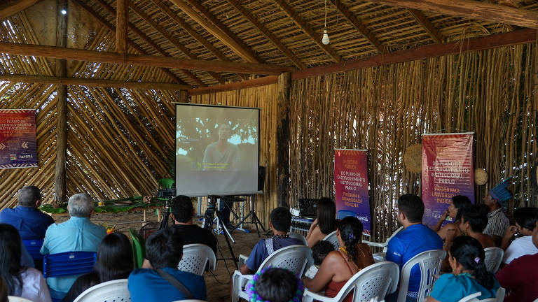 A imagem mostra um grupo de pessoas assistindo a uma apresentação em um espaço coberto com estrutura de palha. Um projetor exibe uma imagem em uma tela grande. As pessoas estão sentadas em cadeiras brancas, e ao fundo há banners coloridos com textos visíveis. O ambiente é rústico e natural, com elementos da cultura local.
