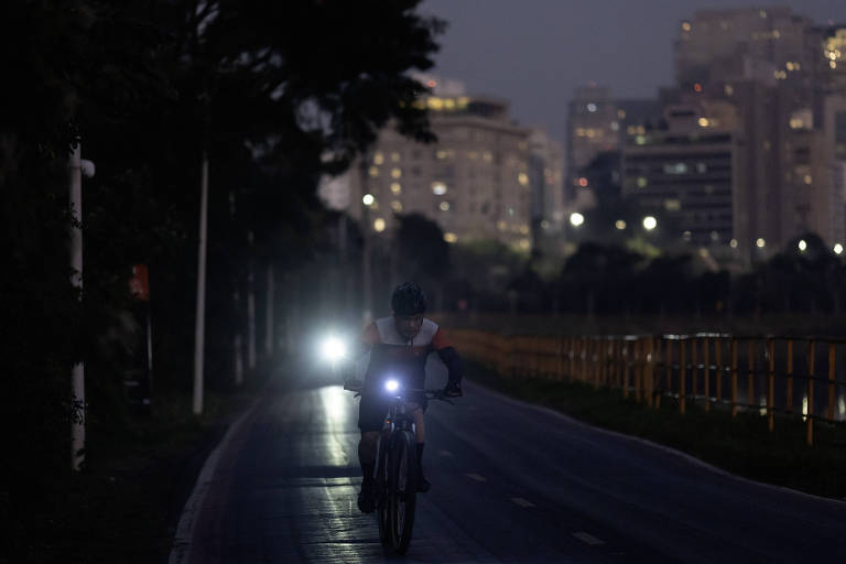 A imagem mostra um ciclista pedalando em uma ciclovia à noite. O ciclista está iluminado por uma luz frontal em sua bicicleta, enquanto ao fundo, há prédios iluminados da cidade. A atmosfera é noturna, com pouca luz natural e algumas luzes artificiais visíveis.