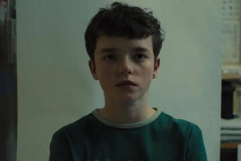 Een jongen met krullend bruin haar staat recht in de camera te kijken. Hij draagt een groen T-shirt met lichte details. De achtergrond is wazig, maar het lijkt een binnenomgeving te zijn, mogelijk een huis, met wat muren en objecten zichtbaar.