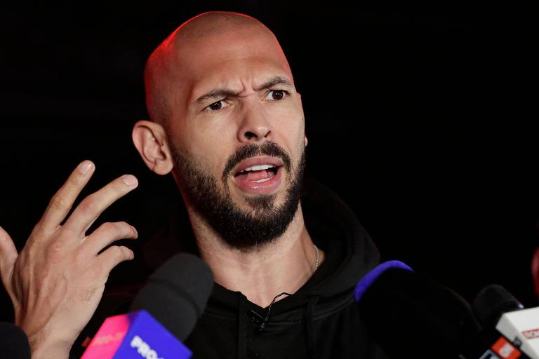 Een man met een kaalgeschoren hoofd en baard is aanwezig bij een persconferentie en gebaart met zijn handen terwijl hij spreekt. Hij draagt een zwart T-shirt en wordt omringd door microfoons van verschillende televisiezenders. Op de achtergrond is het silhouet van een andere persoon te zien, die aandachtig lijkt te luisteren.