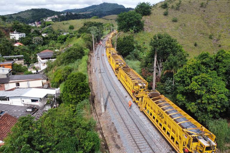 A imagem mostra um trem de manutenção amarelo percorrendo uma linha férrea em uma área rural. À esquerda, há casas e vegetação, enquanto à direita, a paisagem é composta por colinas e árvores. O trem está em movimento, e algumas pessoas podem ser vistas próximas aos trilhos.