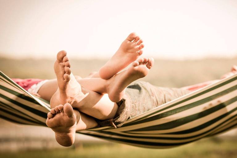 A imagem mostra os pés de duas pessoas relaxando em uma rede. Os pés estão descalços e se entrelaçam, com um fundo desfocado que sugere um ambiente ao ar livre. A luz é suave, criando uma atmosfera tranquila e aconchegante.