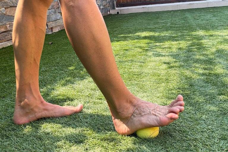 A imagem mostra um pé descalço pressionando uma bola amarela sobre uma superfície de grama sintética. O fundo apresenta uma área externa com uma parede de pedras e uma estrutura de vidro, sugerindo um ambiente de lazer ou exercício. A luz natural ilumina a cena, destacando a textura da grama e a forma do pé.