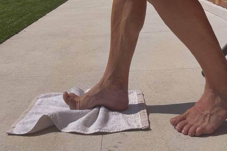 A imagem mostra uma pessoa sentada em uma superfície de concreto, com uma perna dobrada e o pé descalço apoiado em uma toalha. O fundo apresenta um gramado verde e uma construção de tijolos. O céu está parcialmente nublado, com algumas nuvens visíveis.