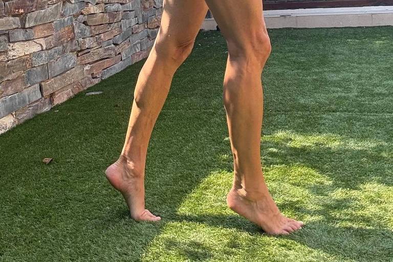 Uma pessoa está caminhando descalça em um gramado verde, com um fundo de parede de pedras e uma casa de madeira ao fundo. A pessoa usa uma camiseta preta e shorts claros, e está em um ambiente ensolarado.