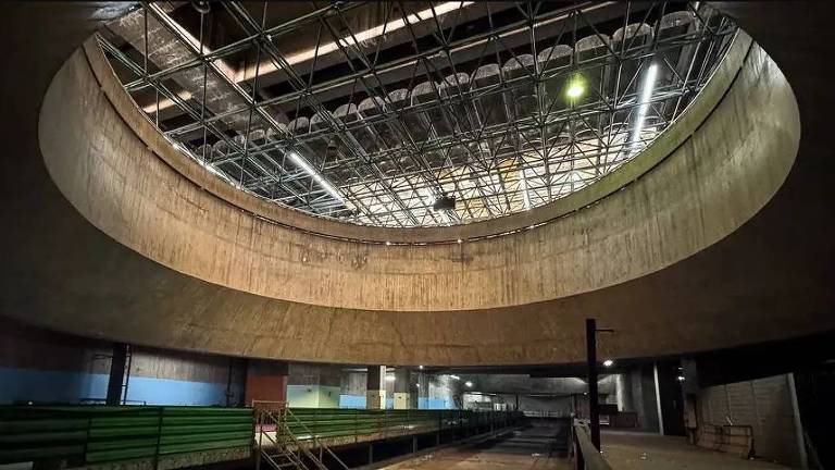 A imagem mostra o interior de uma grande estrutura circular com um teto alto e exposto. O piso é de concreto e há uma iluminação que destaca a forma do teto. As paredes são lisas e de cor clara, e há uma área com bancos ou assentos em um tom verde ao fundo. O ambiente parece ser um espaço amplo e industrial, possivelmente um ginásio ou uma pista de atletismo.