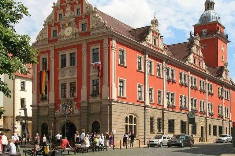 Uma construção histórica de cor vermelha, com detalhes arquitetônicos elaborados, incluindo janelas simétricas e um telhado inclinado. O edifício possui uma torre com um relógio e está decorado com bandeiras. Na frente, há um grupo de pessoas conversando e algumas mesas de café ao ar livre. O céu está parcialmente nublado.