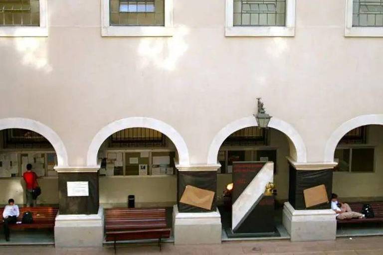 A imagem mostra um pátio de um edifício com arcos. Há bancos de madeira dispostos ao longo do pátio, onde algumas pessoas estão sentadas. As paredes são de cor clara e há janelas com vitrais. O ambiente parece ser bem iluminado, com sombras projetadas no chão. Um dos arcos tem uma placa de aviso ou informação visível.