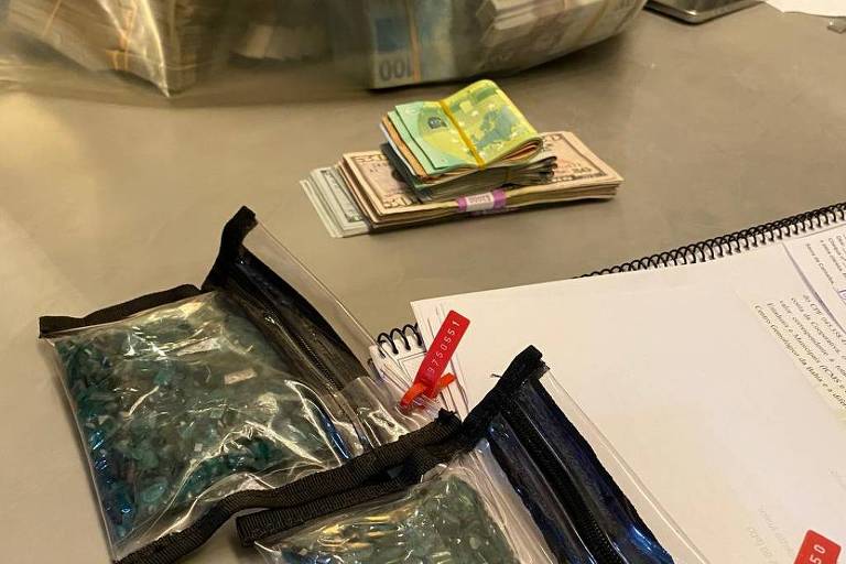 A imagem mostra uma mesa com dois pacotes transparentes contendo objetos não identificáveis, possivelmente pedras ou cristais, e uma pilha de dinheiro em notas ao fundo. Também há uma pequena quantidade de dinheiro em notas visíveis na parte superior da mesa, além de um caderno e um celular.