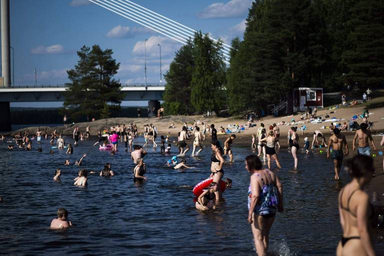 The image shows a beach full of people having fun in the water and on the sand. Many people are swimming and playing in the water, some using floats. In the background, a bridge and surrounding trees can be seen. The sky is clear with a few clouds.