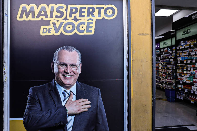 A imagem mostra uma grande placa publicitária com um homem sorridente, vestindo um terno escuro e uma gravata clara. Ele está com a mão sobre o coração e tem óculos. Acima dele, está escrito 'MAIS PERTO DE VOCÊ' em letras grandes e amarelas. Ao fundo, pode-se ver o interior de uma loja com prateleiras cheias de produtos.