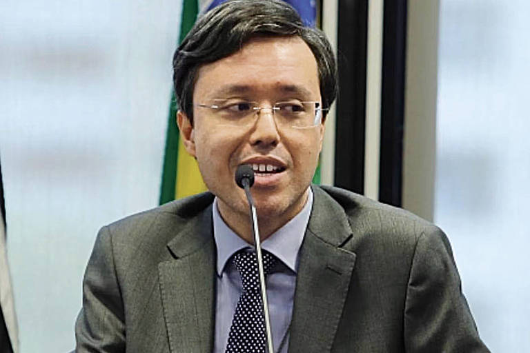 Um homem de terno cinza e gravata, com cabelo liso e escuro, está falando em um microfone. Ele parece estar em um evento ou conferência, com uma mesa à sua frente que tem um copo de água e um nome em um cartão. Ao fundo, há bandeiras do Brasil e de outros países visíveis através de uma janela.