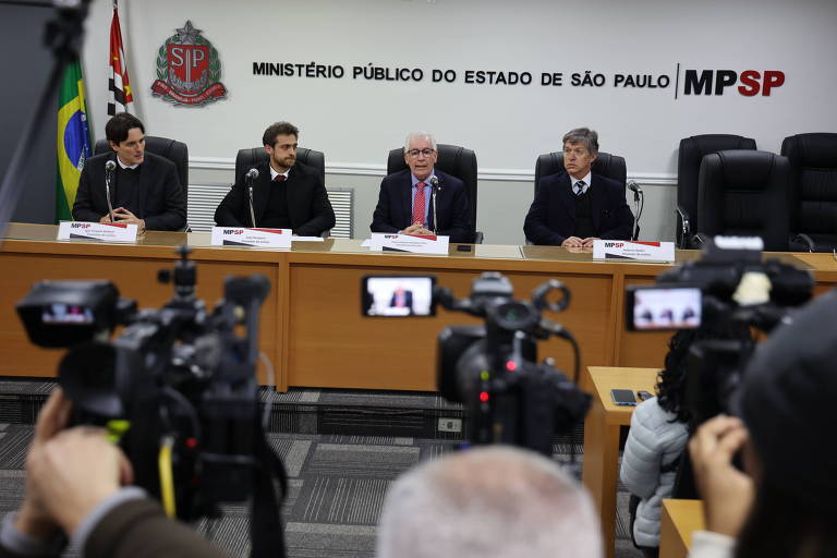 A imagem mostra uma coletiva de imprensa no Ministério Público do Estado de São Paulo. Quatro homens estão sentados em uma mesa, com um deles falando ao microfone. Ao fundo, há bandeiras do Brasil e do estado de São Paulo, além do brasão do Ministério Público. Várias câmeras de jornalistas estão focadas na mesa, indicando que a coletiva está sendo gravada e transmitida.