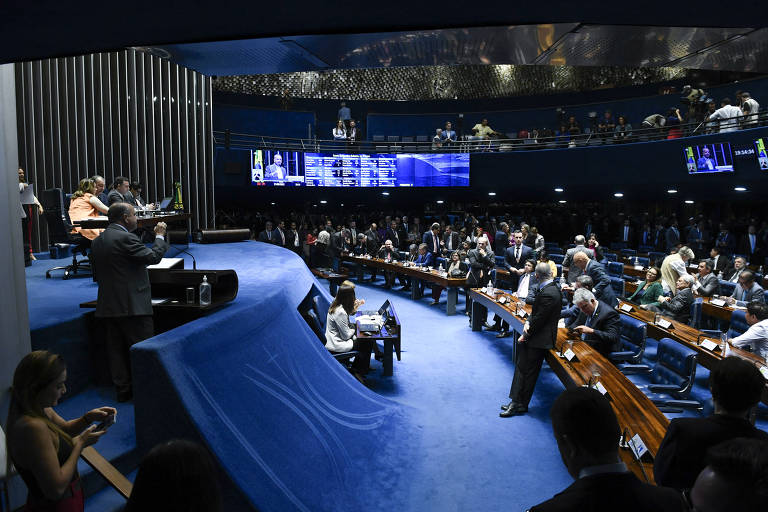 A imagem mostra uma sessão do Senado Brasileiro, com senadores sentados em cadeiras dispostas em um formato semicircular. No centro, um grupo de senadores está em pé, enquanto outros estão sentados em mesas. Ao fundo, há telas exibindo informações, e o ambiente é iluminado com luzes azuis e douradas. O espaço é amplo, com um design moderno e uma grande quantidade de pessoas presentes.