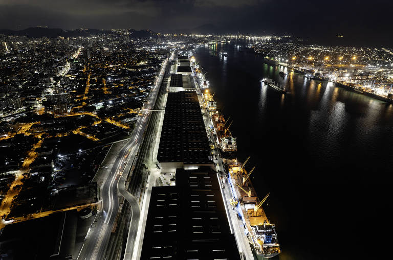 A imagem mostra uma vista aérea noturna de um porto, com armazéns e navios atracados. À esquerda, há uma área urbana iluminada, com ruas e edifícios visíveis. O rio está à direita, refletindo as luzes da cidade e do porto. O céu está escuro, indicando que é de noite.