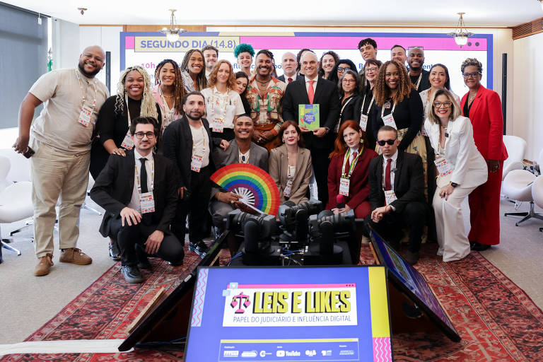 A imagem mostra um grupo grande de pessoas reunidas em um evento. Elas estão posando para a foto em um ambiente interno, com um fundo que inclui uma tela e decoração colorida. Algumas pessoas estão segurando um objeto em forma de arco-íris. A maioria dos participantes está vestida de maneira formal ou semi-formal, e há uma variedade de estilos e etnias entre eles.