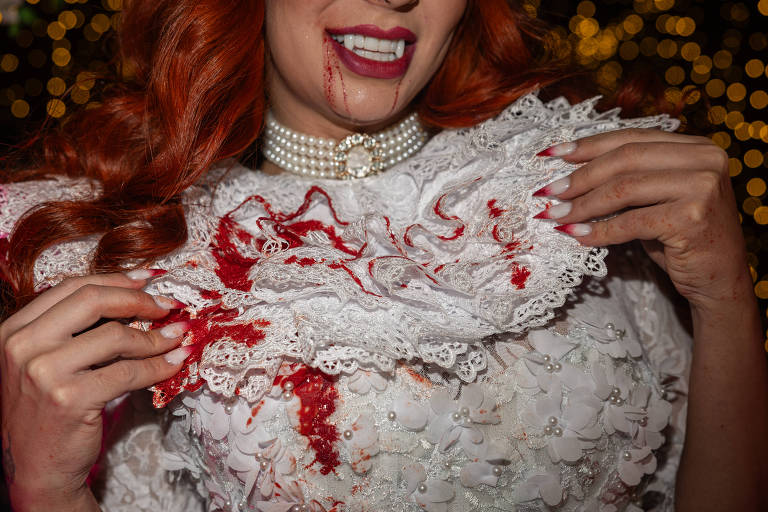 A imagem mostra uma mulher com cabelo ruivo e lábios vermelhos, vestindo um vestido branco decorado com flores e rendas. Ela está segurando a gola do vestido, que apresenta manchas de sangue. O fundo é desfocado com luzes brilhantes, criando um efeito festivo.