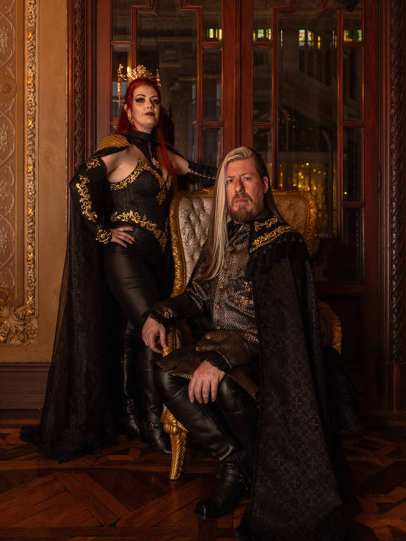 A imagem mostra um casal posando em um ambiente luxuoso. A mulher está em pé, vestindo um traje preto com detalhes dourados e uma coroa, enquanto o homem está sentado em uma cadeira, usando um manto escuro e uma camisa de estilo medieval. O fundo apresenta paredes decoradas e janelas com luz suave, criando uma atmosfera elegante e dramática.