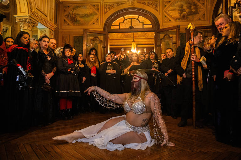 A imagem mostra uma performance em um ambiente gótico, com uma dançarina em destaque, vestindo um top prateado e uma saia branca. Ela está sentada no chão, com uma expressão intensa e uma mão estendida. Ao fundo, há uma multidão de pessoas vestidas em trajes escuros, algumas com acessórios como capas e chapéus. O ambiente é decorado com detalhes arquitetônicos elaborados e iluminação suave, criando uma atmosfera dramática.
