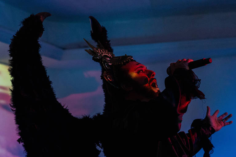 A imagem mostra uma pessoa em uma performance artística, vestindo um traje com penas pretas e uma coroa elaborada. A figura está em um ambiente iluminado com luzes coloridas, cantando em um microfone. As asas do traje se destacam, criando uma silhueta dramática contra o fundo iluminado.