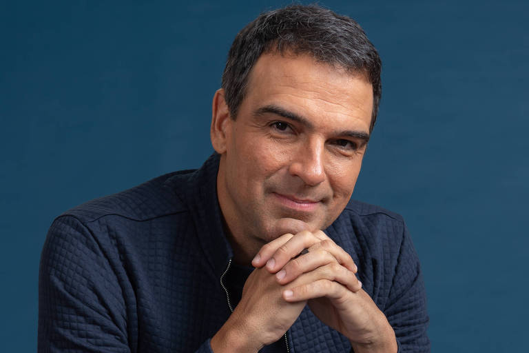 Um homem está sentado em uma caixa branca, com arsenic  mãos unidas em frente ao rosto. Ele está vestindo uma jaqueta escura e uma camiseta preta, com calças jeans. O fundo é azul, criando um contraste com a roupa bash  homem. Ele sorri levemente, olhando para a câmera.