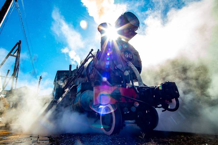 A imagem mostra uma locomotiva a vapor em funcionamento, com vapor saindo de sua chaminé. O sol brilha atrás da locomotiva, criando um efeito de luz intensa. O céu é azul e há algumas nuvens. A locomotiva está posicionada em uma plataforma, e o chão é de pedras. A cena transmite uma sensação de movimento e energia.
