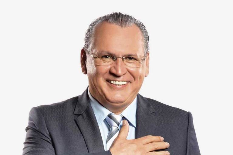Um homem sorridente está posando para a foto. Ele usa um terno escuro, uma camisa clara e uma gravata listrada. O homem está com a mão sobre o coração, em um gesto de saudação ou respeito. O fundo da imagem é branco, destacando a figura do homem.