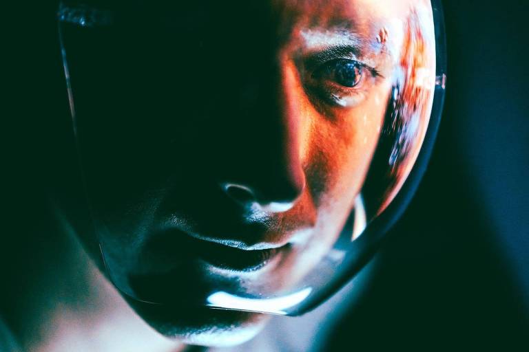 De afbeelding toont een close-up van het gezicht van een astronaut, met een reflecterende helm die zijn gezichtsuitdrukking benadrukt. De belichting is dramatisch, met tinten blauw en oranje die een opvallend contrast vormen. De achtergrond is donker, waardoor het figuur van de astronaut en de glans van de helm worden benadrukt.