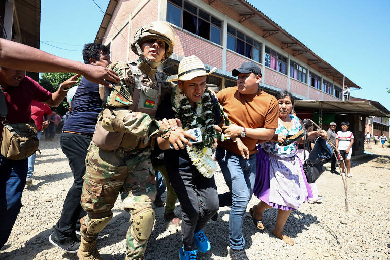 De afbeelding toont een groep mensen in een conflictsituatie. Een soldaat, gekleed in militair uniform en helm, houdt een man vast die van de plaats delict lijkt te worden verwijderd. De man draagt een hoed en een T-shirt en lijkt zich in een weerstandspositie te bevinden. Om hem heen bewegen andere mensen snel, sommigen met een bezorgde blik. De setting is een landelijk gebied met een gebouw op de achtergrond en de grond bedekt met rotsen.
