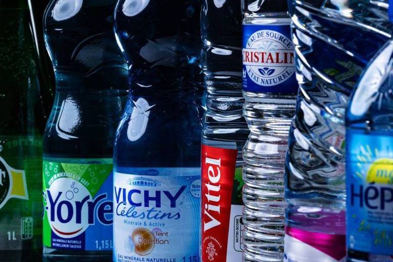 A imagem mostra uma variedade de garrafas de água dispostas em uma linha. As garrafas incluem marcas como Perrier, Yorre, Vichy Celestins, Vittel, Cristalit e Hepar. As garrafas têm diferentes formatos e tamanhos, com algumas sendo transparentes e outras com rótulos coloridos. O fundo é escuro, destacando as garrafas.