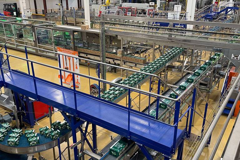 A imagem mostra uma visão panorâmica de uma fábrica com uma linha de produção. Há várias esteiras transportadoras em funcionamento, com produtos em caixas verdes sendo movidos. Estruturas metálicas e plataformas azuis estão presentes, além de equipamentos e maquinários ao fundo. O ambiente é bem iluminado e organizado, com um piso amarelo.