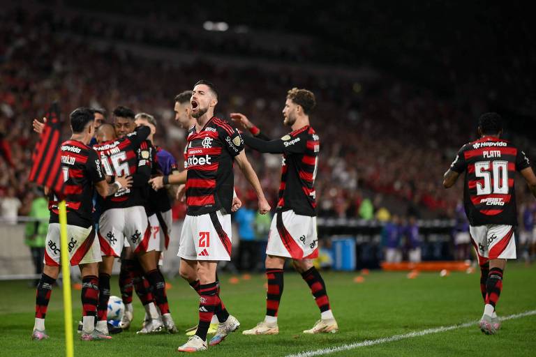 Um grupo de jogadores do Flamengo celebra um gol em um estádio lotado. Os jogadores estão vestidos com uniformes listrados em vermelho e preto, e alguns deles estão se abraçando e comemorando. O ambiente é vibrante, com torcedores ao fundo, muitos deles segurando bandeiras e fazendo barulho.
