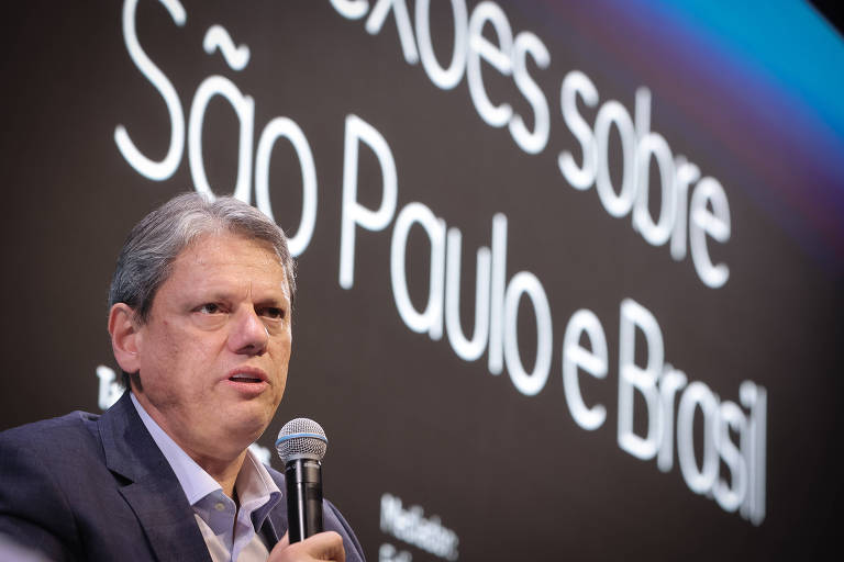 Um homem com cabelo grisalho e vestindo um paletó escuro está falando ao microfone. Ao fundo, há um painel com o texto "Reflexões sobre São Paulo e Brasil" em letras grandes e brancas. O ambiente parece ser um auditório ou uma sala de conferências.