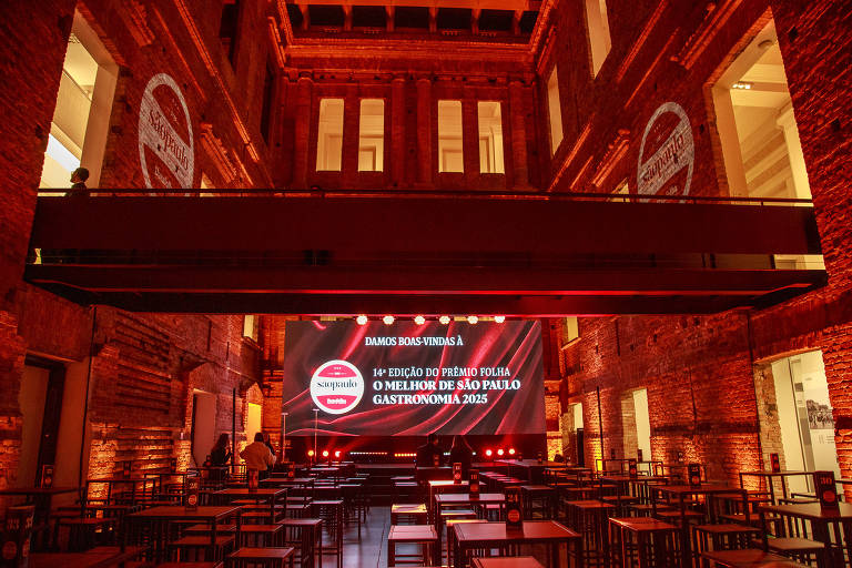 A imagem mostra um espaço interno de um evento, com paredes de tijolos expostos e iluminação em tons quentes. No centro, há um palco com um grande telão exibindo a mensagem: '10ª EDIÇÃO DO PRÊMIO FOLHA O MELHOR DE SÃO PAULO GASTRONOMIA 2023'. O ambiente está decorado com mesas e cadeiras dispostas para os convidados, e há uma atmosfera de expectativa e celebração.