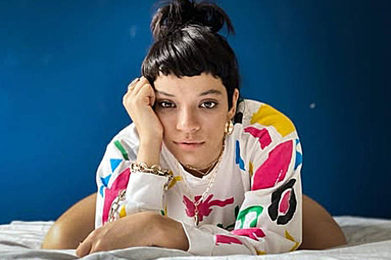A imagem mostra uma jovem mulher deitada sobre uma cama, com a cabeça apoiada na mão. Ela tem cabelo curto e escuro, e usa uma blusa colorida com padrões variados. O fundo da imagem é uma parede azul, e a cama é coberta com um lençol claro. A expressão dela é pensativa e relaxada.