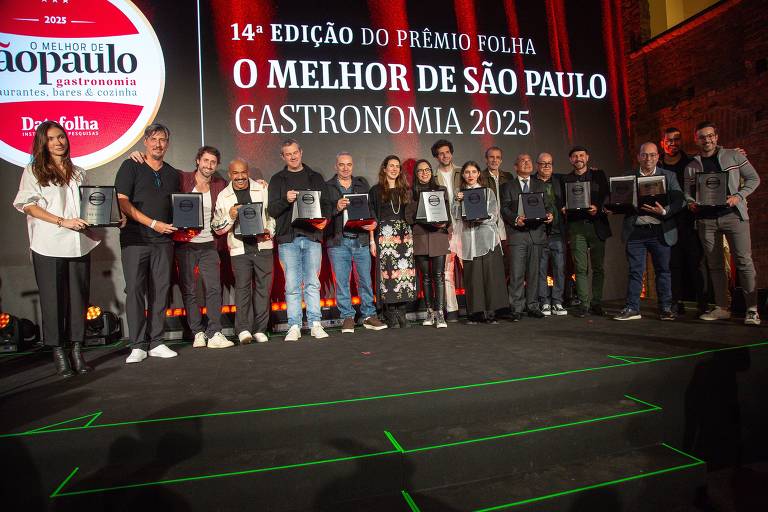 A imagem mostra um grupo de pessoas em um evento de premiação, posando em um palco. Eles estão segurando placas de reconhecimento. Ao fundo, há um grande banner com o texto '14ª EDIÇÃO DO PRÊMIO FOLHA O MELHOR DE SÃO PAULO GASTRONOMIA 2025'. A iluminação é suave e o ambiente parece festivo.