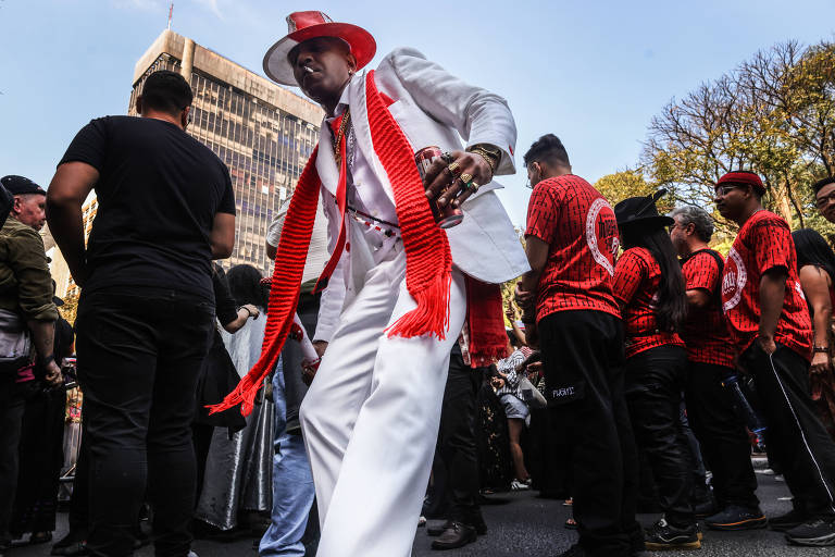 A imagem mostra um grupo de pessoas em um desfile de Carnaval. Em primeiro plano, um homem vestido com um terno branco e um chapéu vermelho, segurando um copo. Ele está dançando e usando um cachecol vermelho. Ao fundo, há várias pessoas vestidas com camisetas vermelhas e pretas, algumas com cabelo longo e outros com cabelo curto. O cenário é urbano, com prédios e árvores ao fundo.