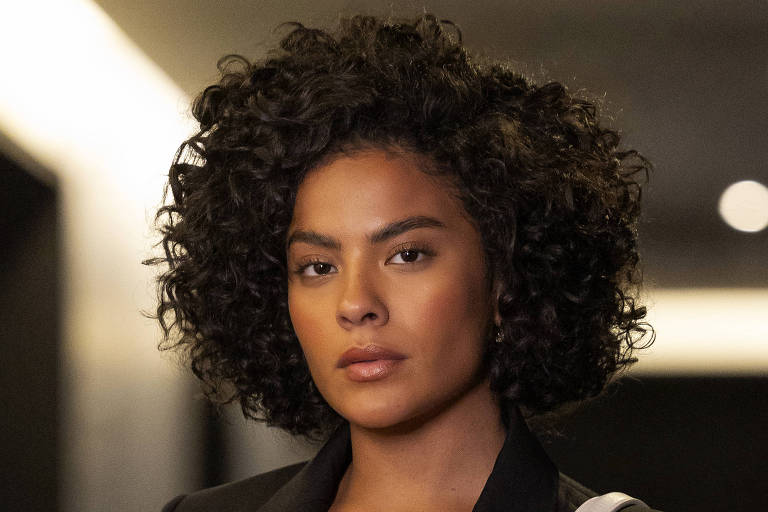 Uma mulher com cabelo cacheado e escuro está caminhando por um corredor iluminado. Ela usa um blazer preto com botões dourados e calças pretas justas. Em uma das mãos, ela segura uma bolsa branca. O corredor tem paredes claras e luzes embutidas nary  teto, criando um ambiente moderno.