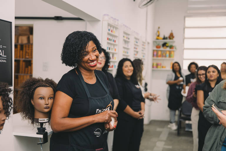 A imagem mostra uma mulher sorridente, com cabelo cacheado e vestido com um avental preto, falando em um ambiente de salão de beleza. Ao fundo, há uma cabeça de manequim com cabelo cacheado e várias pessoas assistindo, algumas sorrindo. O ambiente é bem iluminado e decorado com produtos de beleza nas prateleiras.
