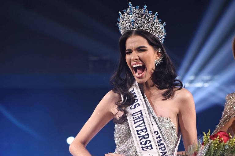 A imagem mostra uma mulher sorridente, vestindo um vestido brilhante e uma coroa, segurando um buquê de flores e uma faixa com os dizeres 'Miss Universo Brasil'. Ao fundo, outras participantes do concurso estão visíveis, vestindo vestidos coloridos. A mulher parece estar celebrando sua vitória.