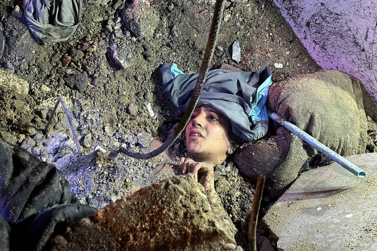 A imagem mostra uma pessoa parcialmente enterrada sob escombros e terra, com a cabeça visível. A pessoa está usando uma roupa escura e um capuz azul. Ao redor, há pedras, sujeira e outros detritos, indicando um cenário de deslizamento ou colapso. A iluminação parece ser artificial, destacando a situação de emergência.