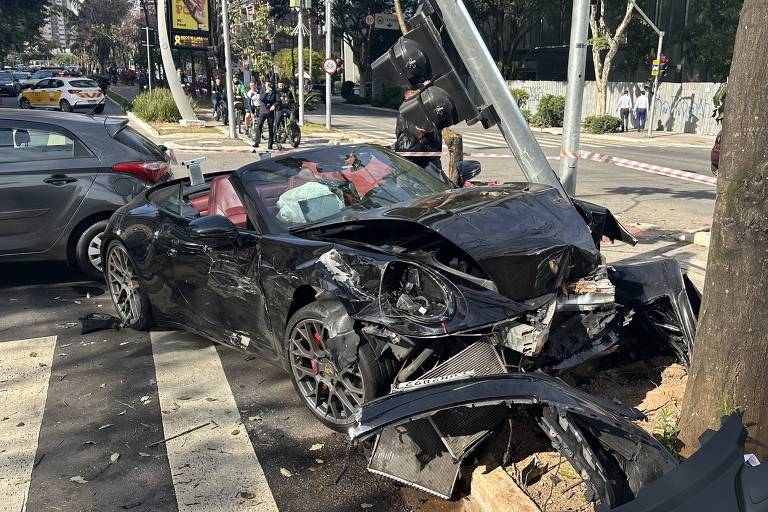 De afbeelding toont een zwarte sportwagen met aanzienlijke schade aan de voorkant, die tegen een boom op de stoep is gebotst. De auto heeft een rood interieur en bevindt zich in een stedelijke omgeving, met andere voertuigen op de achtergrond. Er zijn mensen in de buurt, mogelijk getuigen van het ongeval, en er zijn verkeerslichten zichtbaar.
