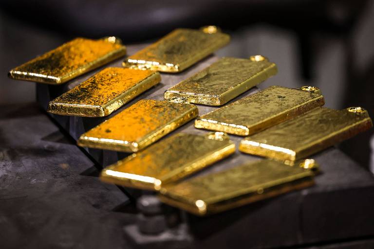 A imagem mostra vários lingotes de ouro dispostos em uma superfície escura. Os lingotes têm um brilho metálico e algumas marcas visíveis em suas superfícies, indicando que foram manuseados. A iluminação destaca o brilho e a cor dourada dos lingotes, criando um contraste com o fundo.