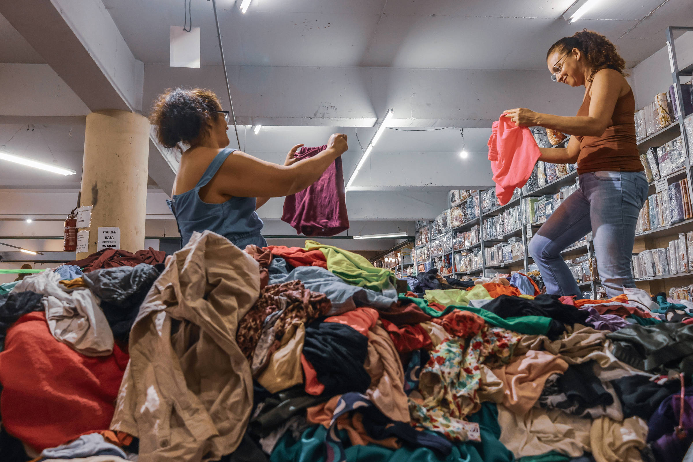 Lojas de roupas por quilo atraem clientes, que sobem em pilhas para garimpar; veja vídeo